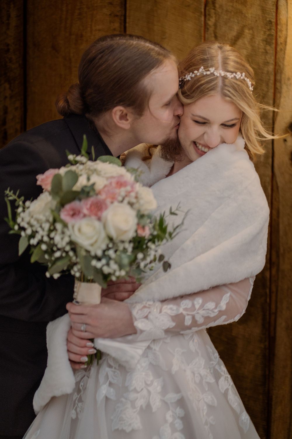 Groom kissing bride on cheek as she smiles, holding pink and white bouquet, wearing white lace gown with floral headpiece.