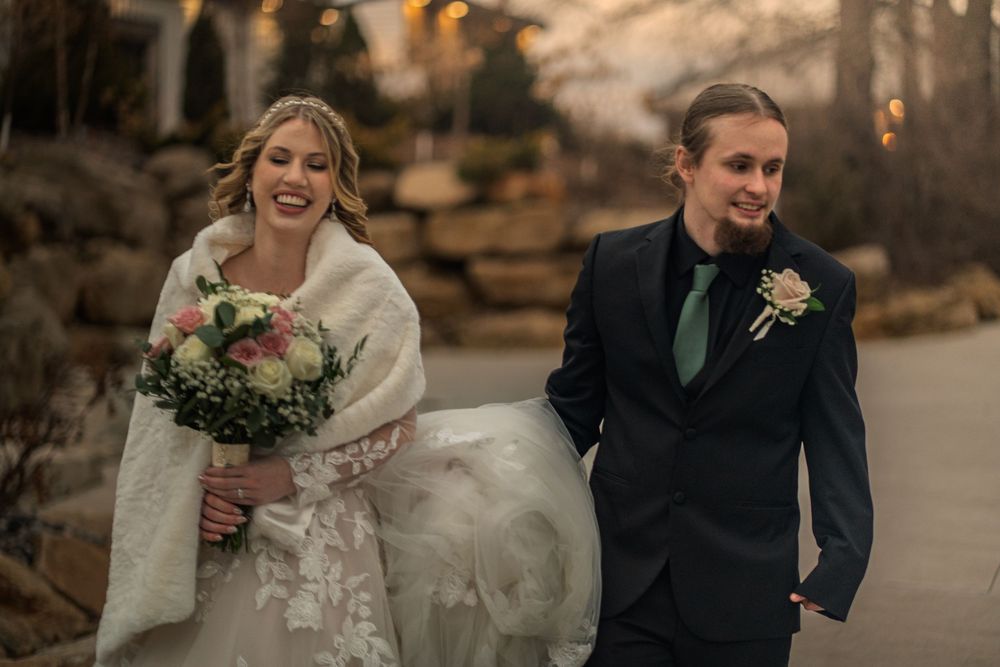 Bride in lace gown with fur wrap holding bouquet walks beside groom in navy suit with green tie at outdoor winter wedding.
