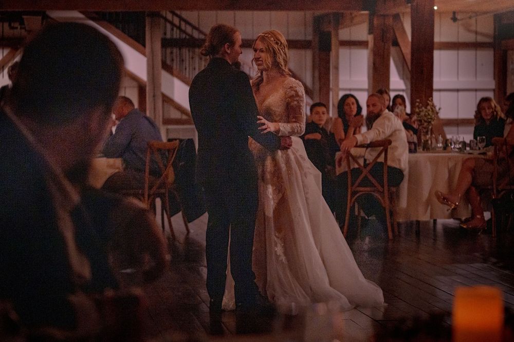 Bride and groom share their first dance in a warmly lit rustic barn venue with wedding guests seated in the background.