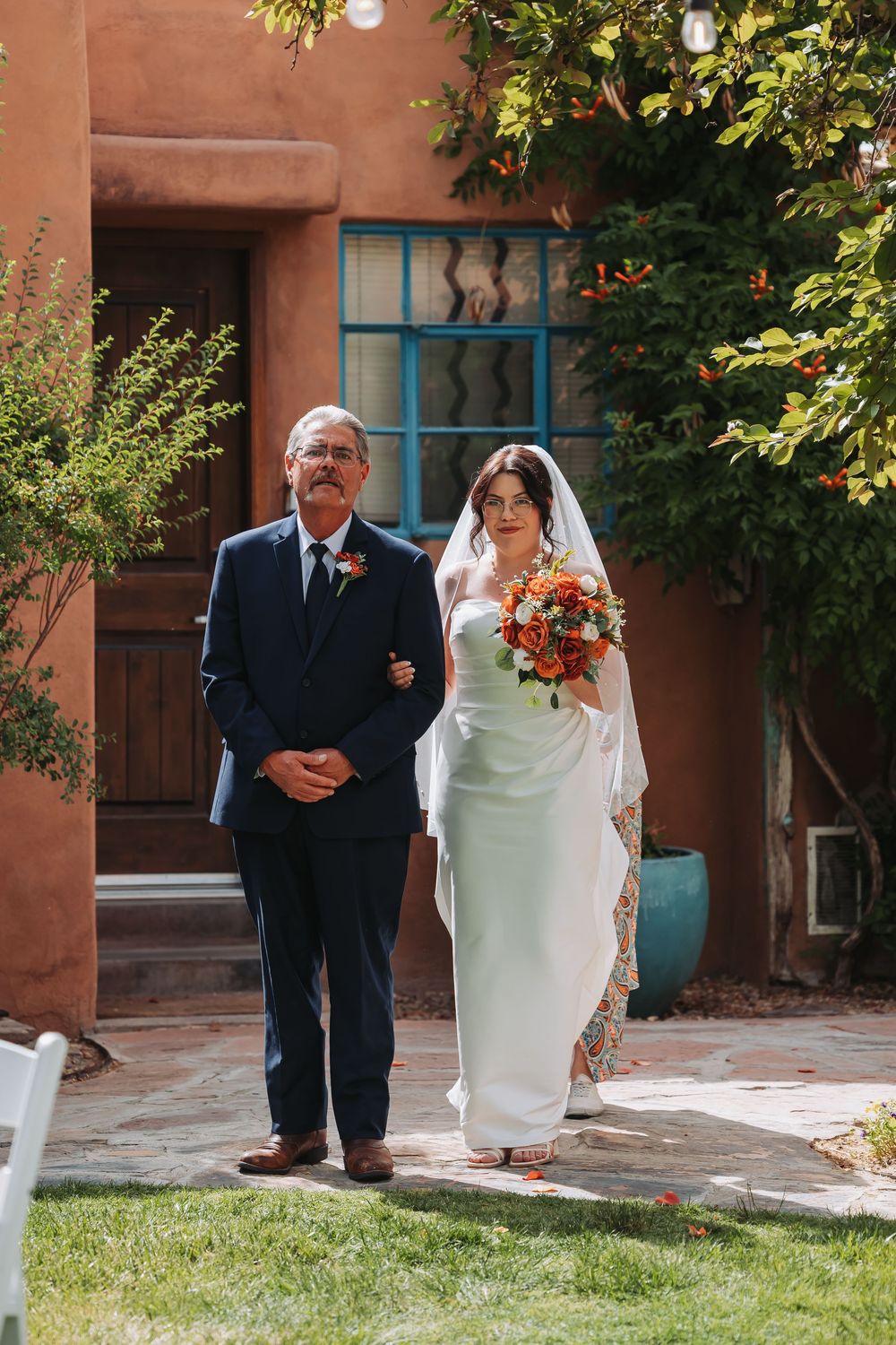 Bride in white gown with veil holding orange bouquet walks with suited man past adobe building.