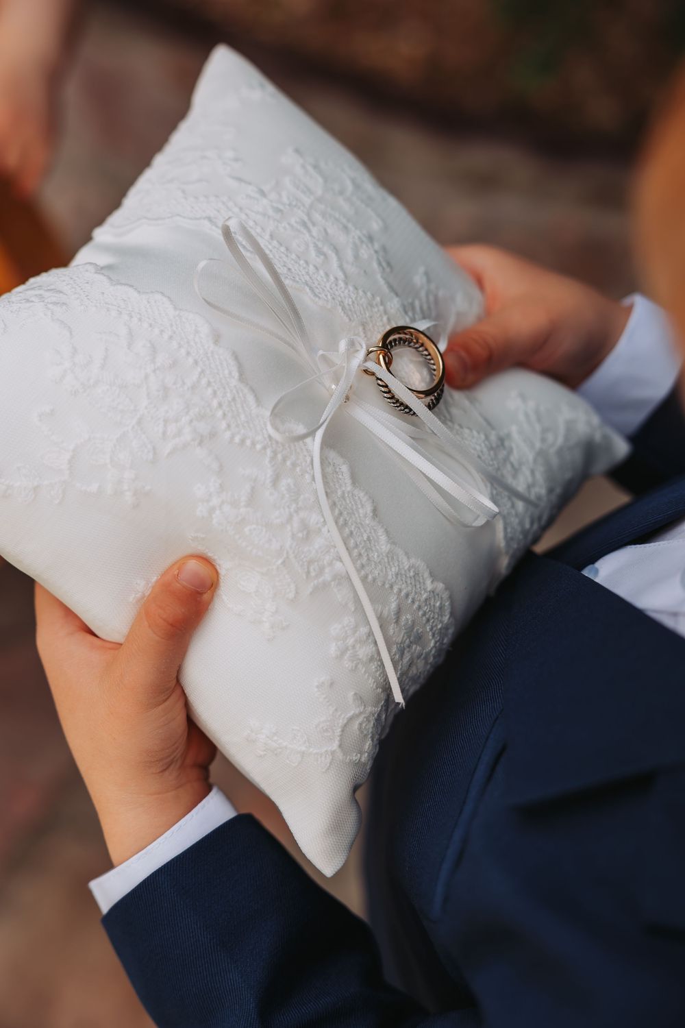 Child in navy suit holding white lace ring bearer pillow with gold wedding bands tied with ribbon bow.