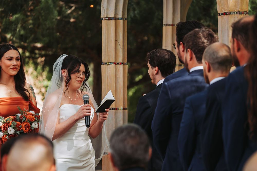 Bride in white gown reads vows at outdoor wedding ceremony, with bridesmaids in orange and groomsmen in navy suits.