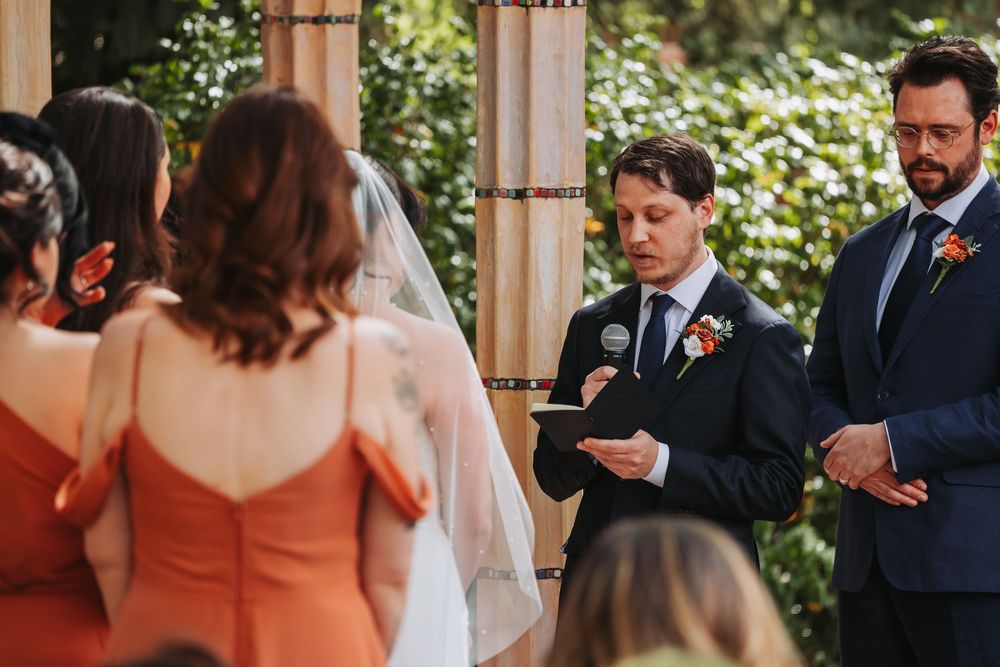 Groom reads vows at outdoor wedding ceremony with bridesmaid in orange dress and best man in navy suit.