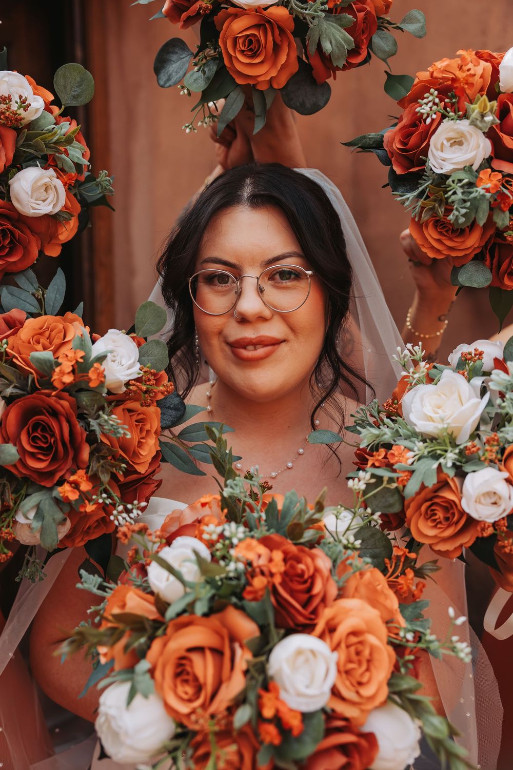 Bride surrounded by vibrant orange and white rose bouquets, wearing glasses and a veil on her wedding day.