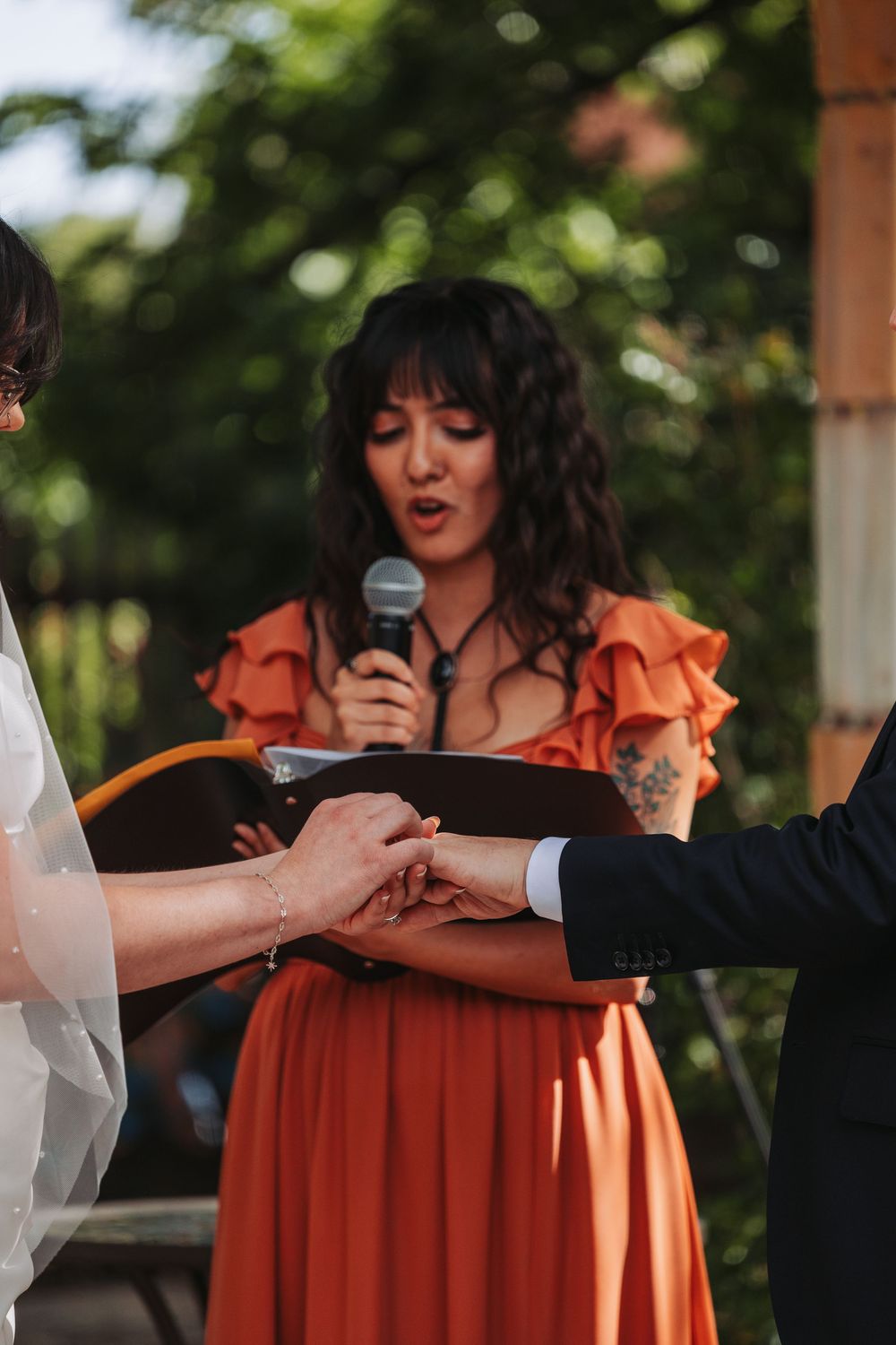 Wedding officiant in orange dress reads vows while holding hands of bride and groom at outdoor ceremony.