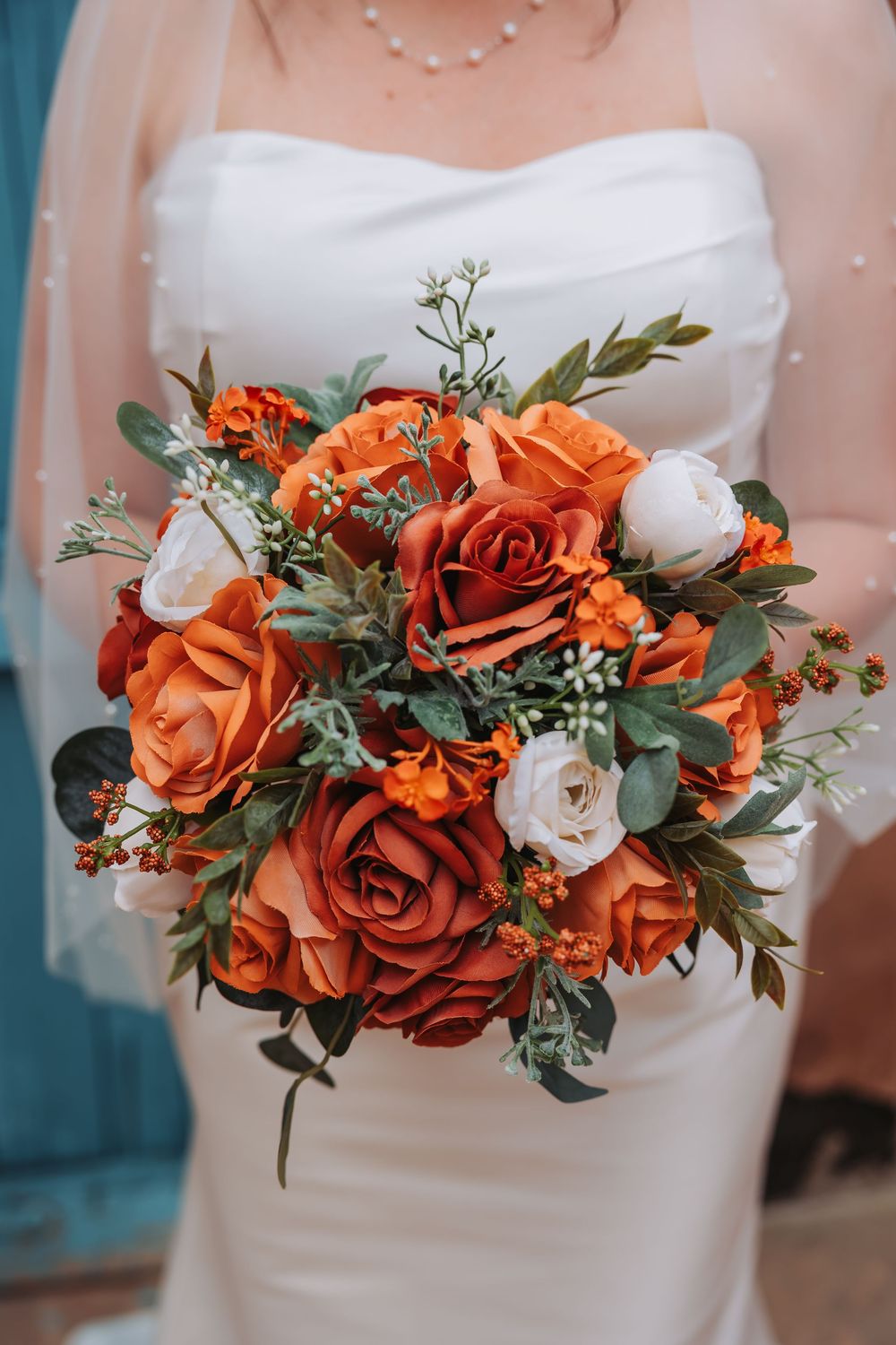Bride in white strapless gown holding a vibrant bouquet of orange, rust, and white roses with greenery.