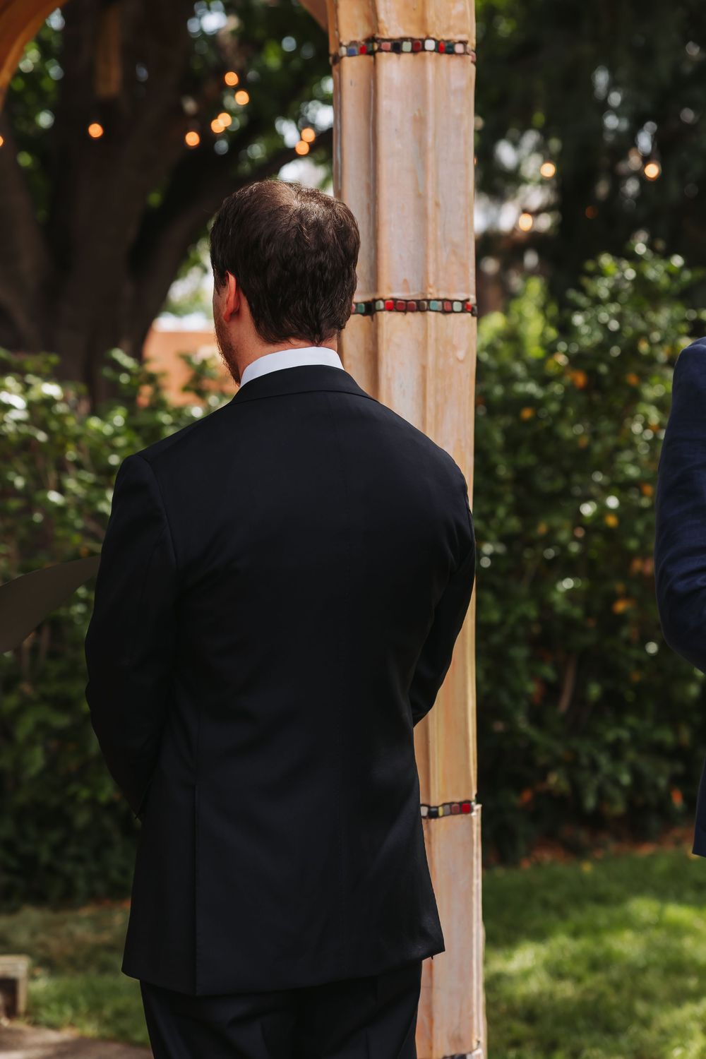 Man in black suit standing at outdoor wedding ceremony altar decorated with colorful beads.