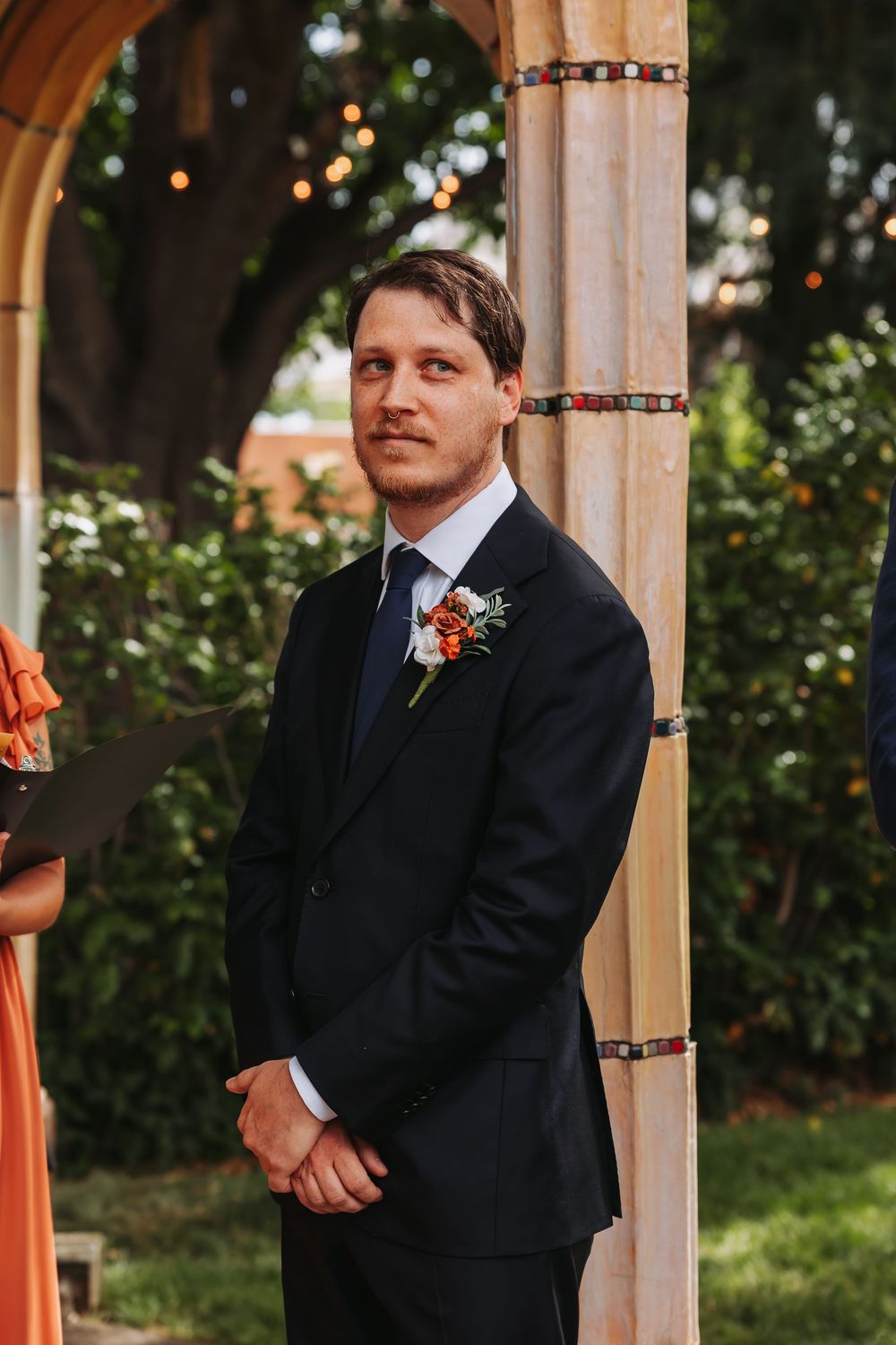 Groom in black suit with floral boutonniere stands at outdoor wedding altar, smiling with anticipation.