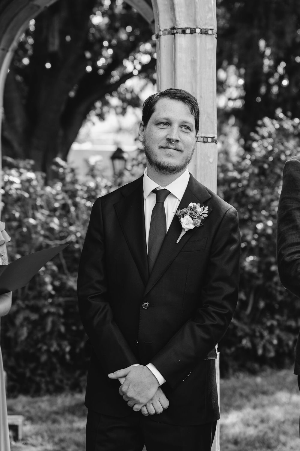 Groom in black suit with boutonniere smiling at wedding ceremony outdoors in black and white photo.