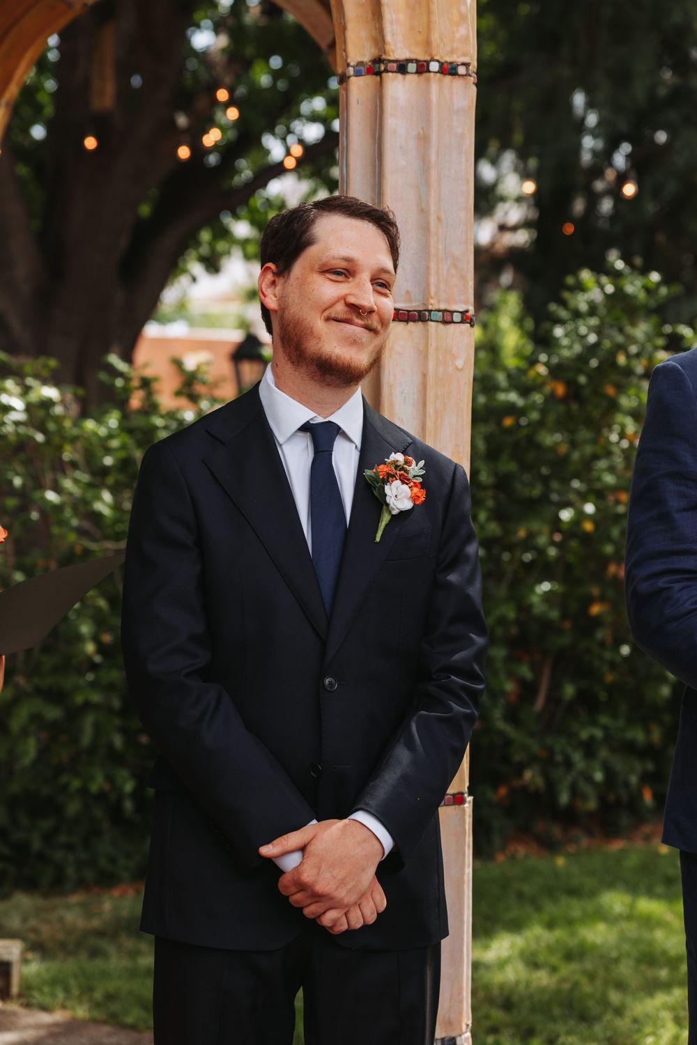 Groom in black suit with boutonniere smiling at outdoor wedding ceremony near wooden arch.