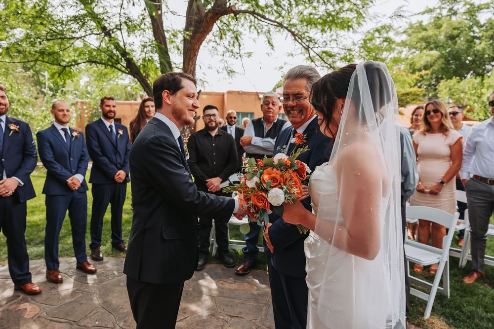 Outdoor wedding ceremony with groom shaking hands with father of the bride holding orange bouquet, guests in background.