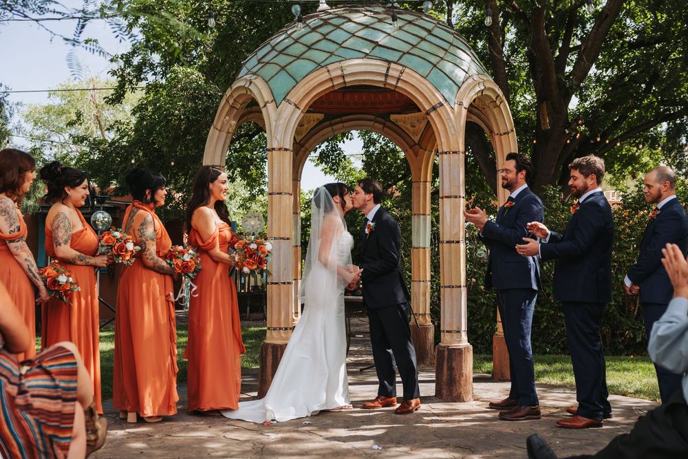 Bride and groom exchange vows under ornate arch with bridesmaids in orange dresses and groomsmen at outdoor wedding ceremony.