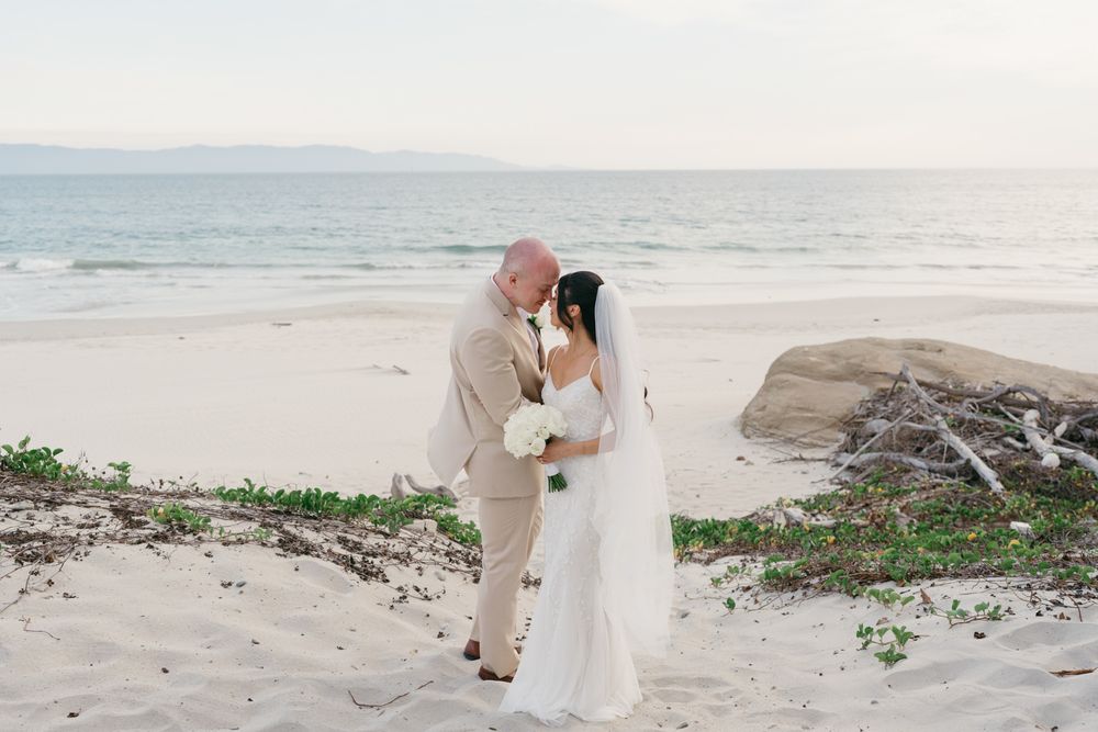 Bride and groom share a romantic kiss on a sandy beach at sunset, with ocean waves in the background.