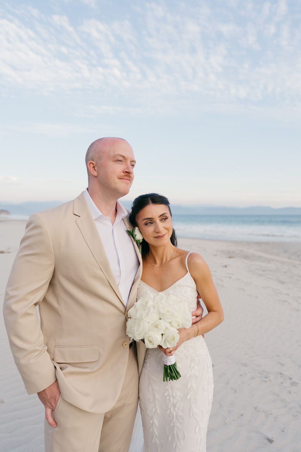 Bride and groom posing on a sandy beach, bride holding white bouquet, groom in tan suit, ocean backdrop.