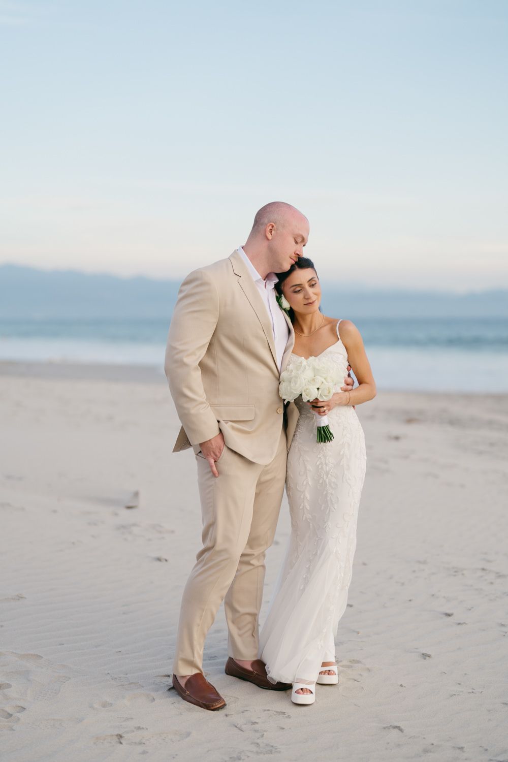 Bride and groom share a tender moment on a sandy beach, she holds white bouquet, he wears tan suit.