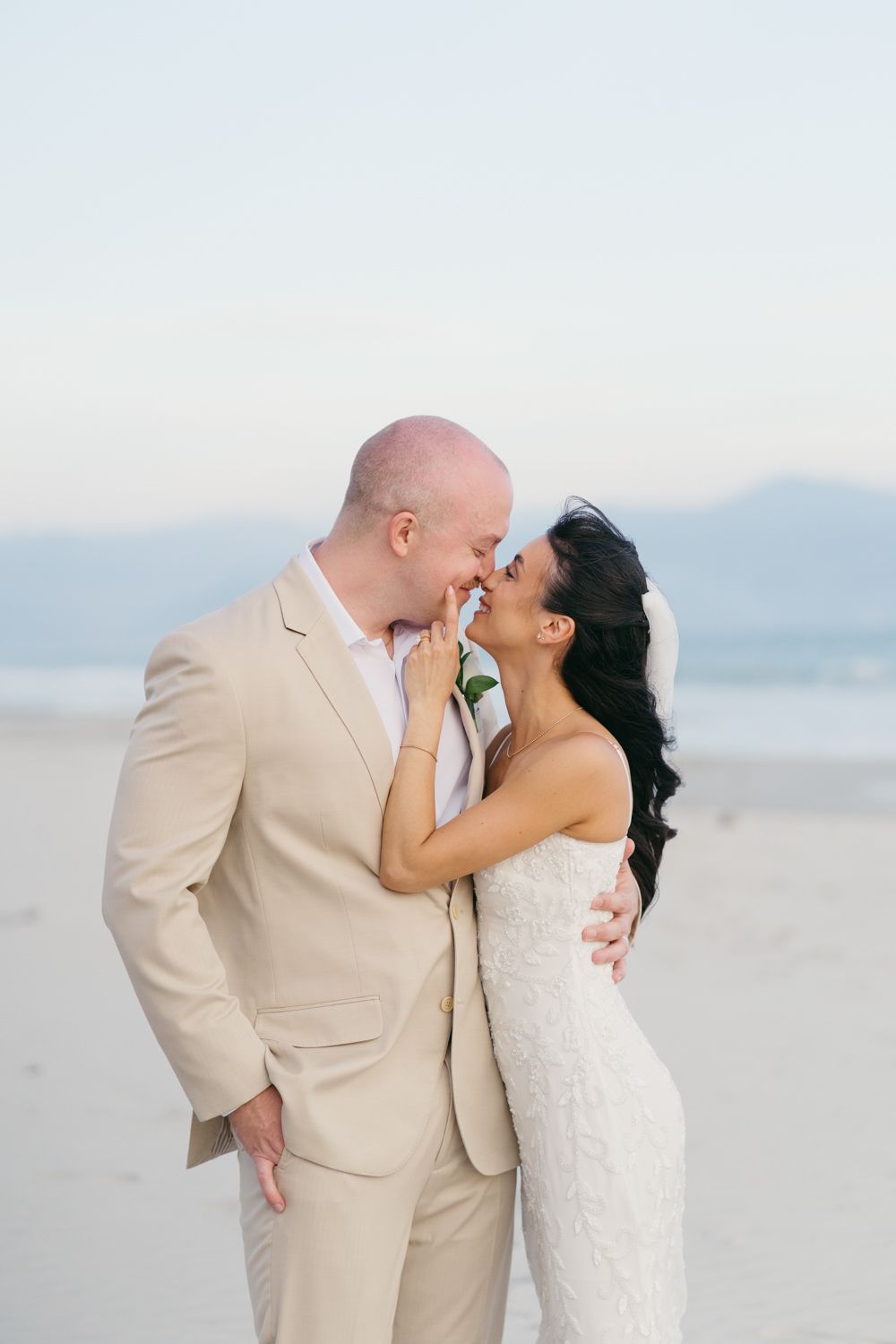 Couple in wedding attire sharing an intimate moment on a serene beach, groom in tan suit, bride in white gown.
