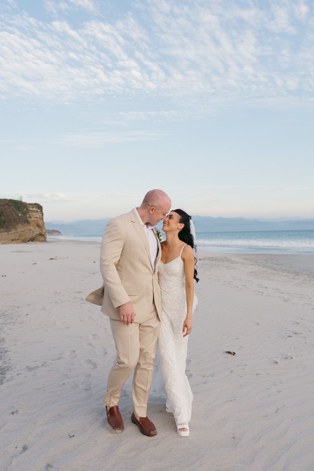 Bride and groom share a romantic moment on a sandy beach, with the groom in a tan suit and bride in a white gown.