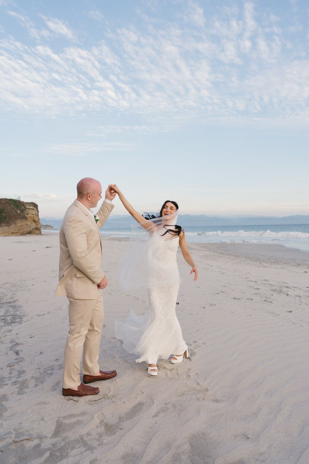 A bride and groom dance joyfully on a sandy beach, with the ocean and blue sky in the background.
