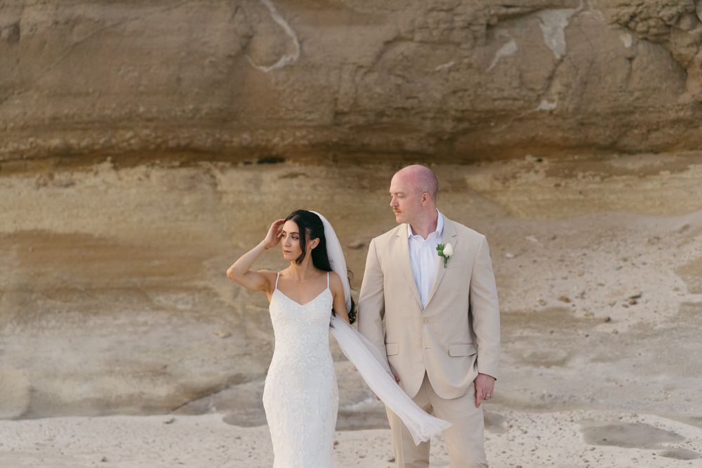 A bride in a white gown and groom in a tan suit walk together on a sandy beach with rocky cliffs behind them.