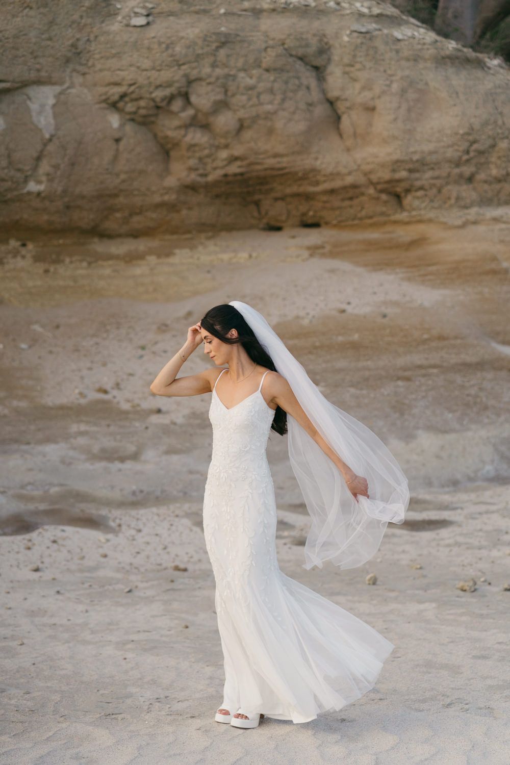 Elegant bride in a fitted white lace gown and flowing veil poses gracefully on a sandy beach near rocky cliffs.