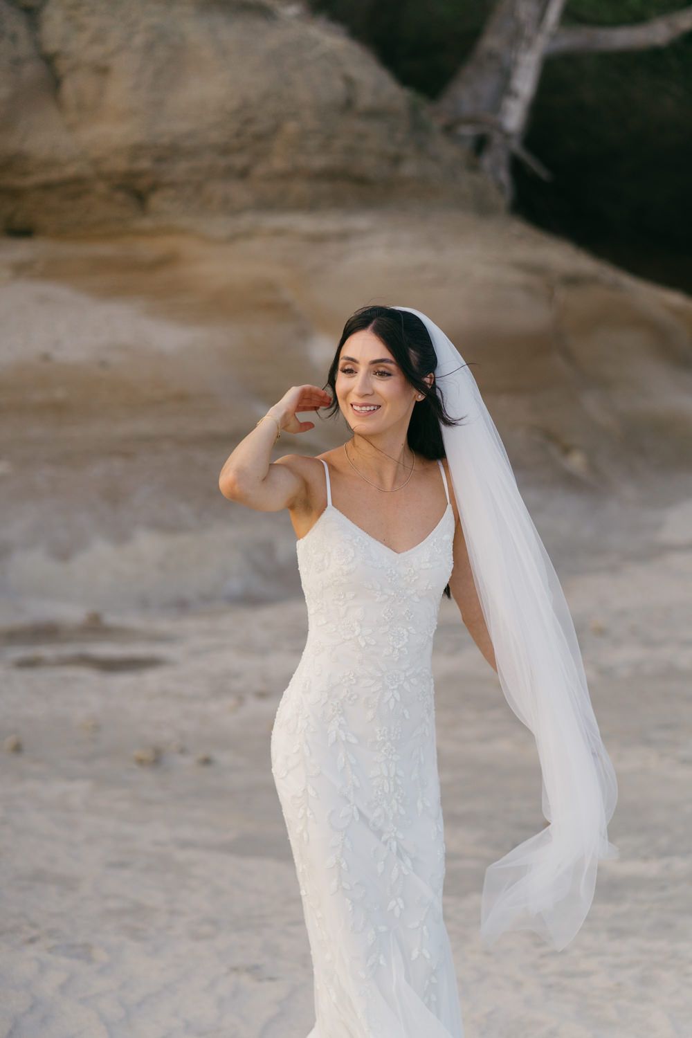 Bride in white lace gown and veil smiles on sandy beach, with rocky cliffs in the background.