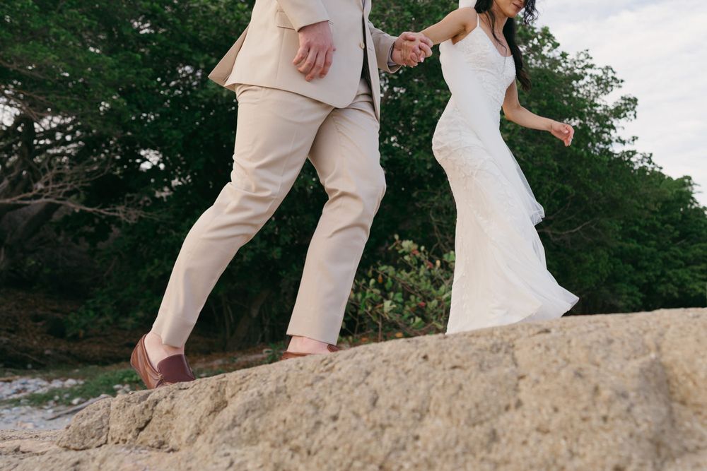 Bride in white gown and groom in tan suit walk hand in hand across rocky coastal terrain.