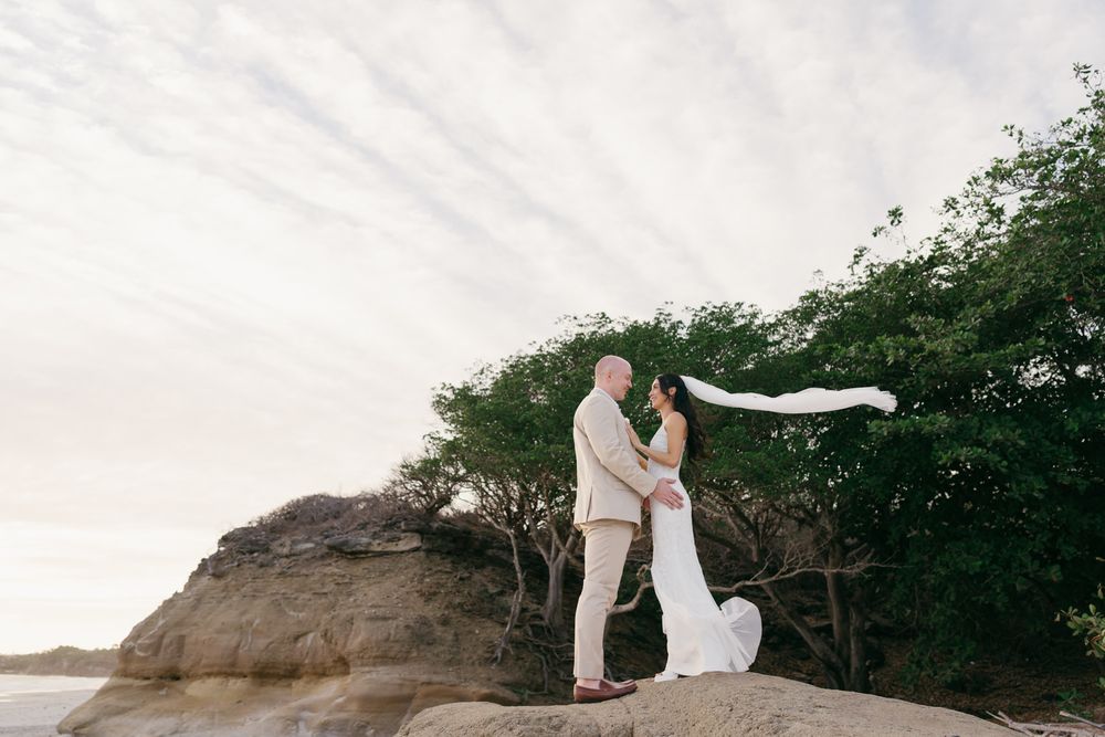 Bride and groom stand on rocky coastal cliffs, bride's veil flowing in wind, lush green trees behind them.