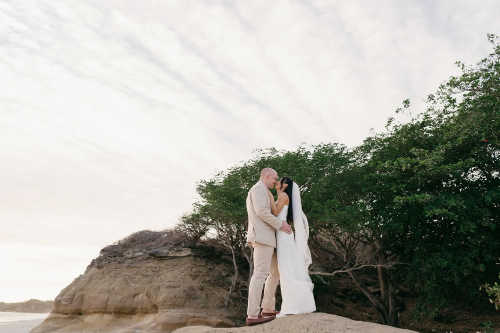 Newlyweds share a romantic kiss on rocky seaside cliff surrounded by lush green trees.