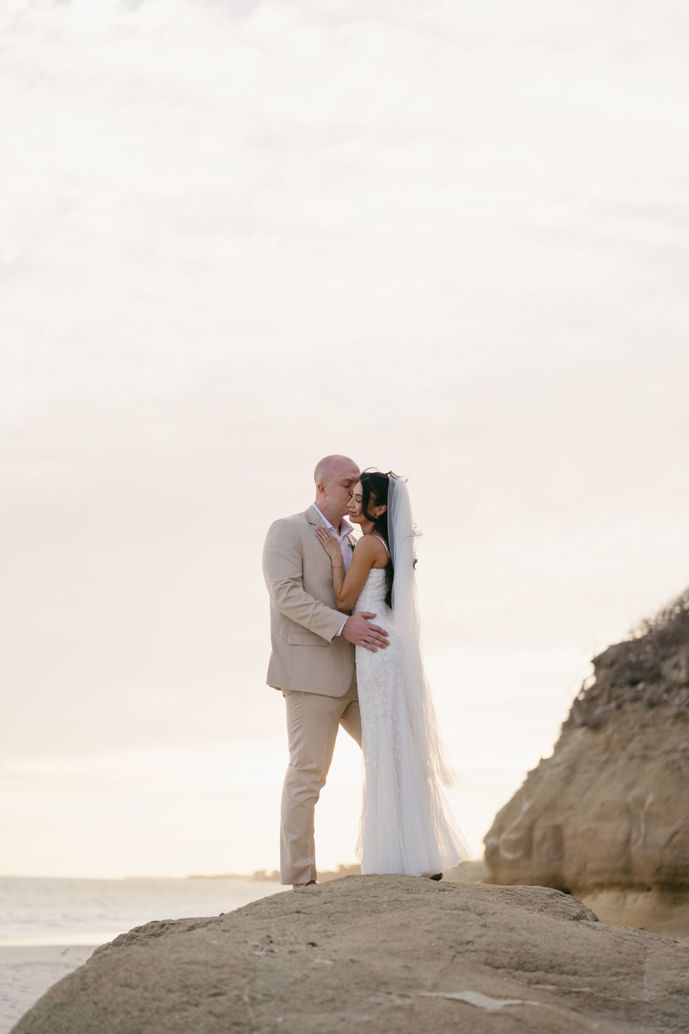 Bride and groom embrace on rocky beach at sunset, groom in tan suit, bride in white gown with veil.