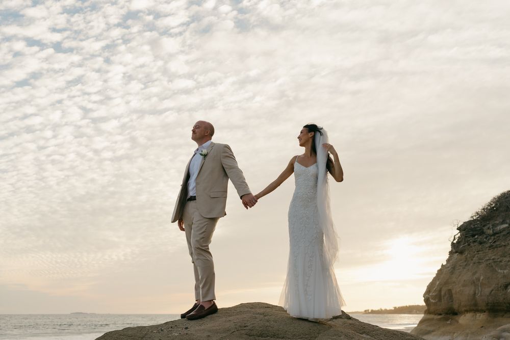 Bride and groom holding hands on coastal rocks at sunset, wearing elegant white gown and tan suit.