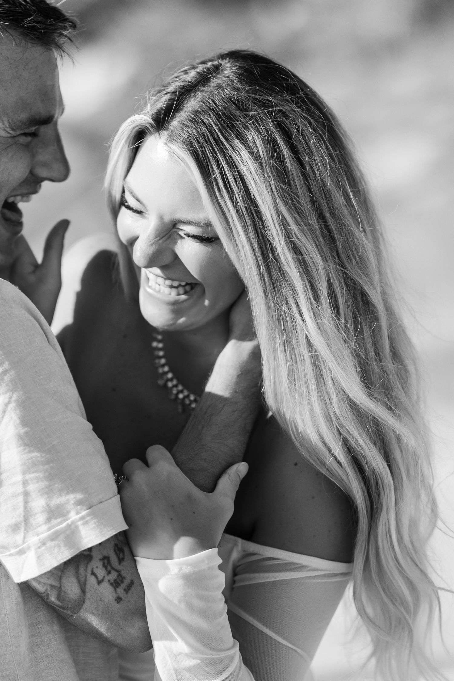 Black and white photo of a laughing blonde woman embracing a tattooed man outdoors in bright sunlight.