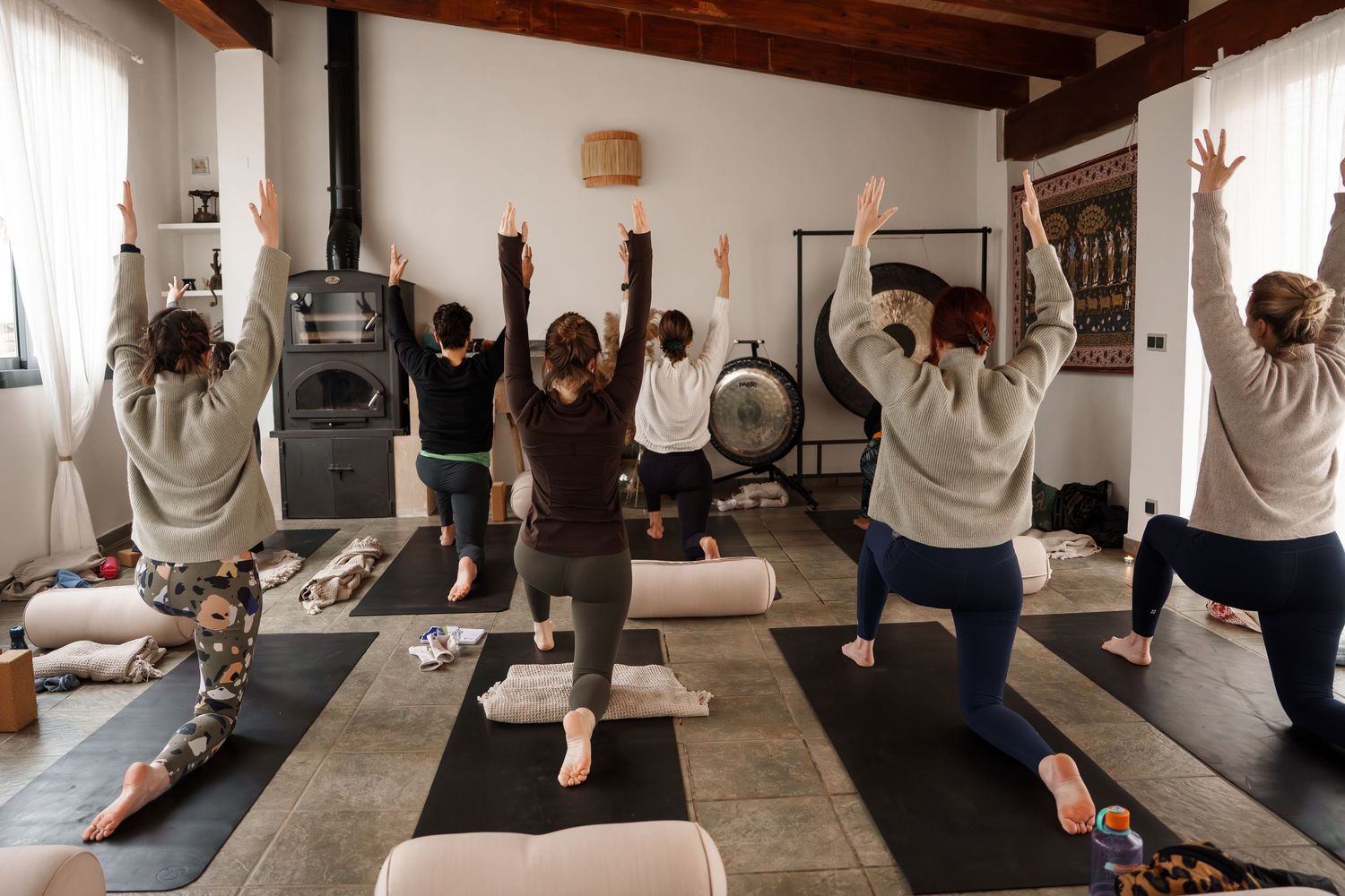 Group yoga class practicing warrior pose with arms raised in a cozy studio with wood stove and natural light.