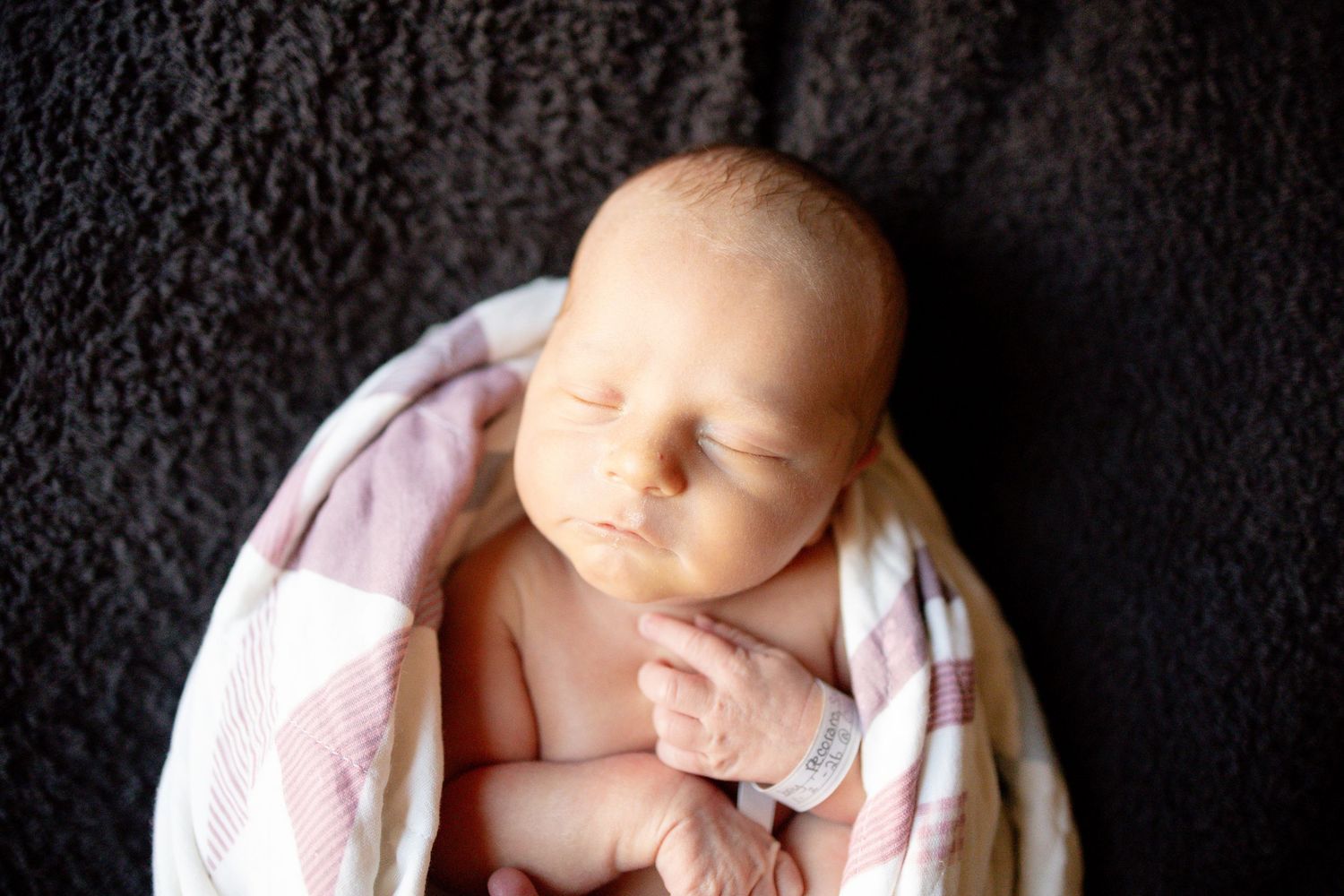 Peaceful newborn baby swaddled in pink and white blanket sleeping on dark gray background.