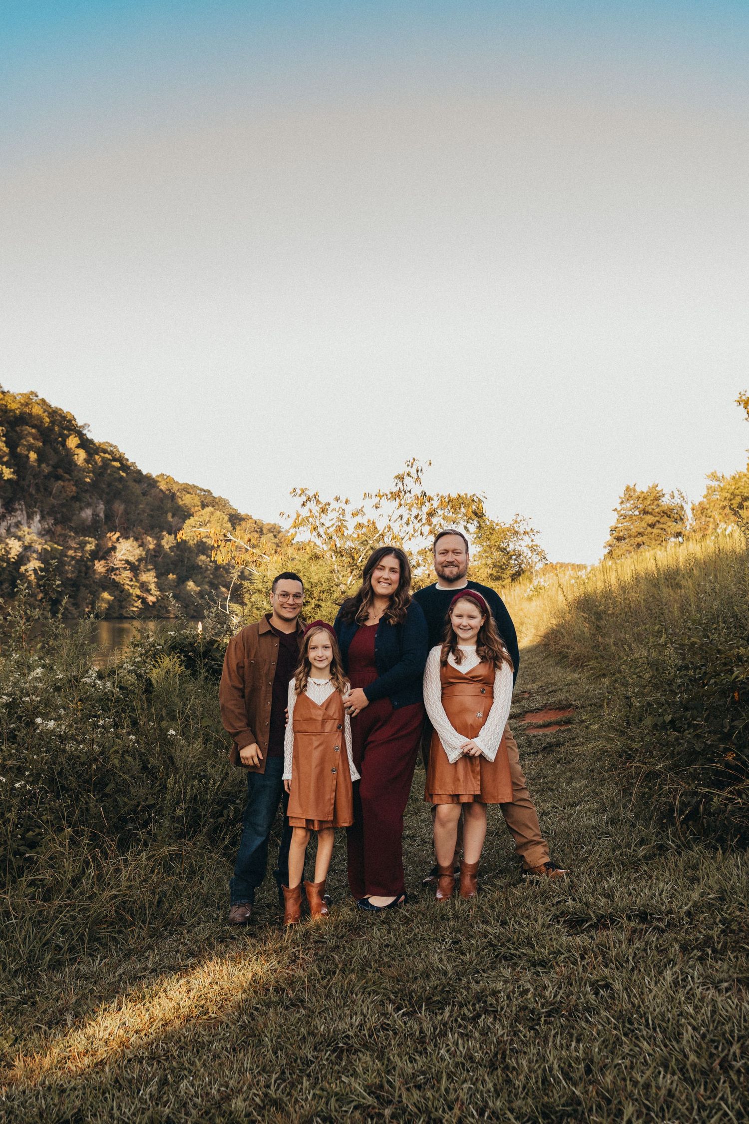 Family of five posing outdoors in autumn attire on a grassy hillside with trees and a river in the background.