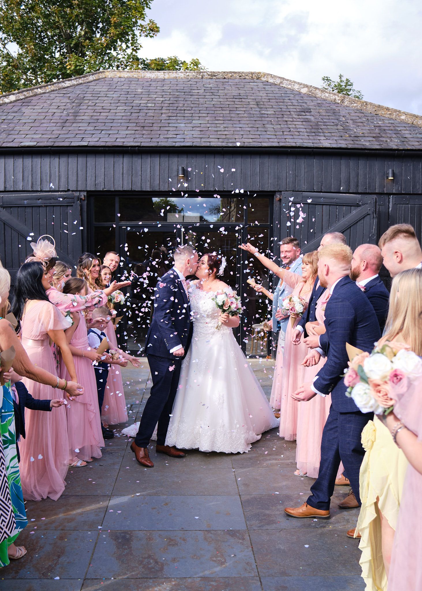 Bride and groom kissing during a colorful confetti shower outside a black barn wedding venue.