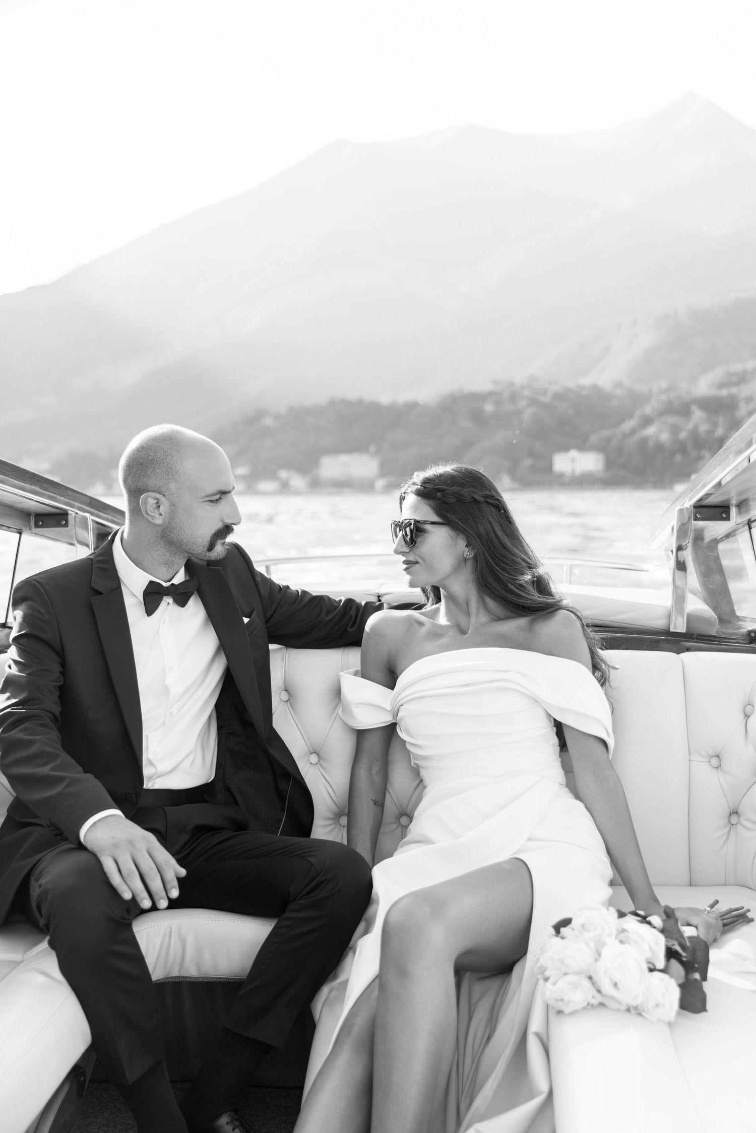Romantic boat ride featuring newlyweds in formal attire against a mountainous lake backdrop.