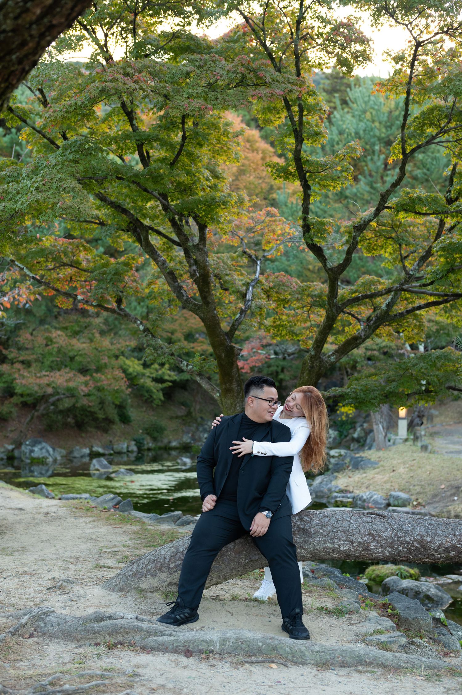 Couple posing affectionately on rocks beside a tranquil Japanese garden pond.