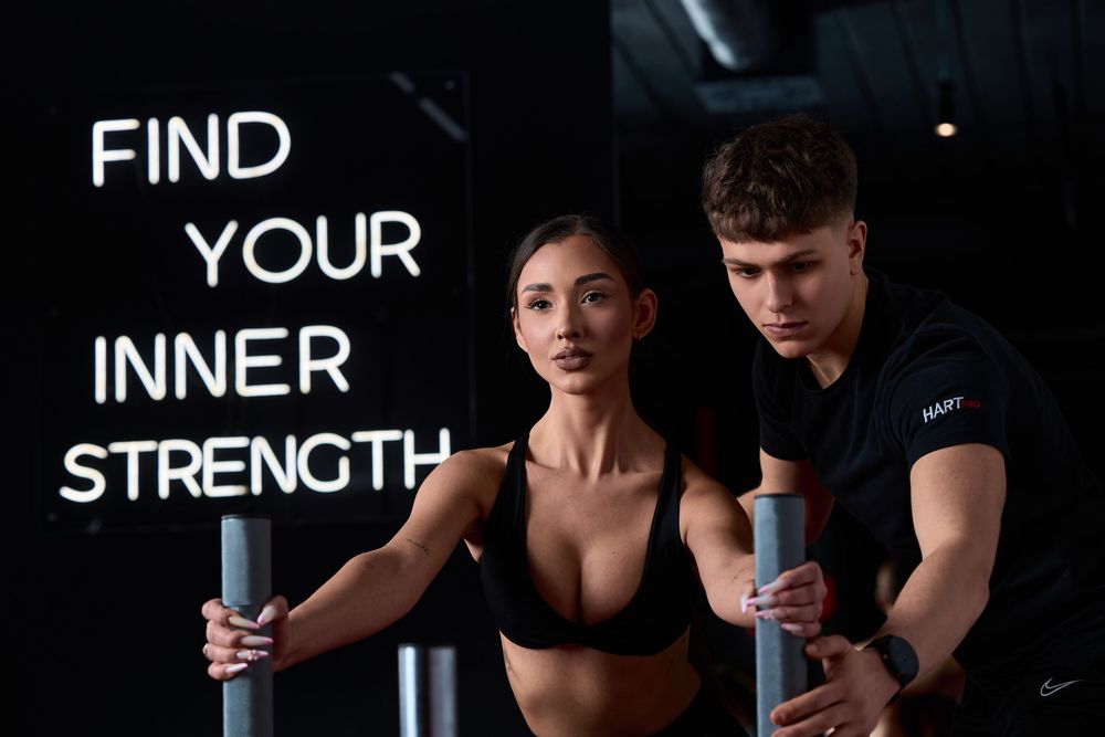 A man and woman grip parallel bars in a dark gym with 'Find Your Inner Strength' neon sign behind them.