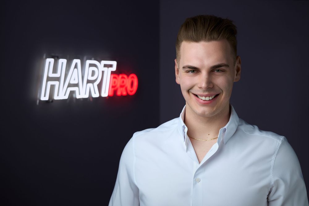 Young man smiling in front of a HartPro neon sign, wearing a white shirt against a dark background.