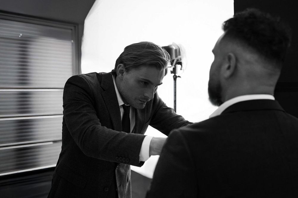 Two men in dark suits, one adjusting the other's sleeve in a black and white professional photo.