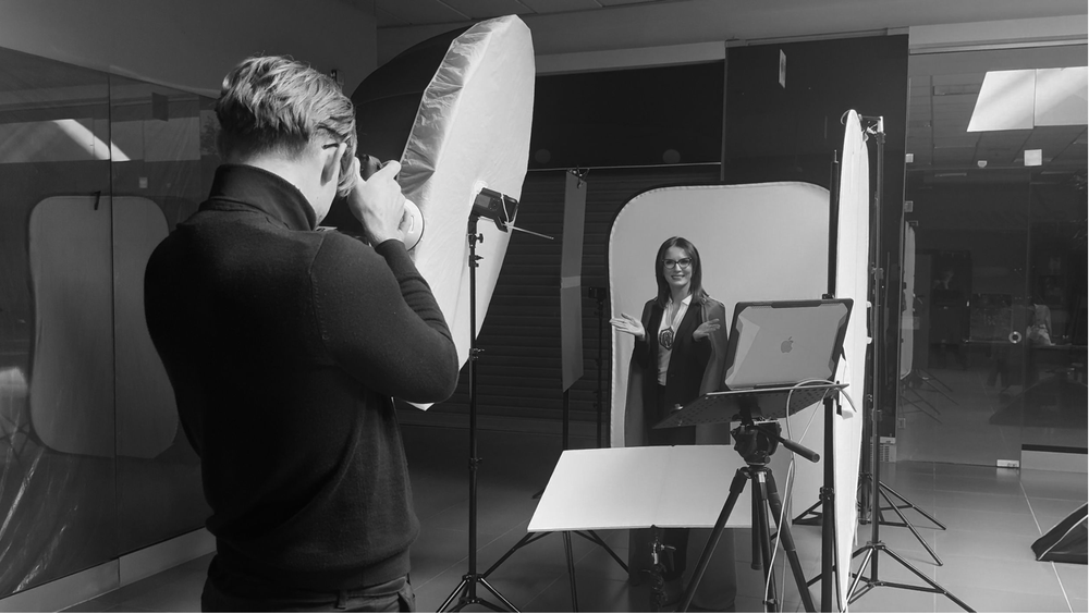 Black and white photo of a photographer taking a professional headshot of a woman in a studio setting.