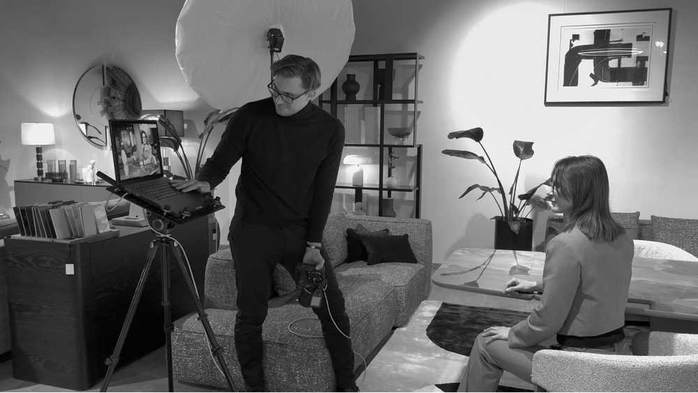 Black and white photo of a photographer filming a woman sitting on a sofa in a modern living room.