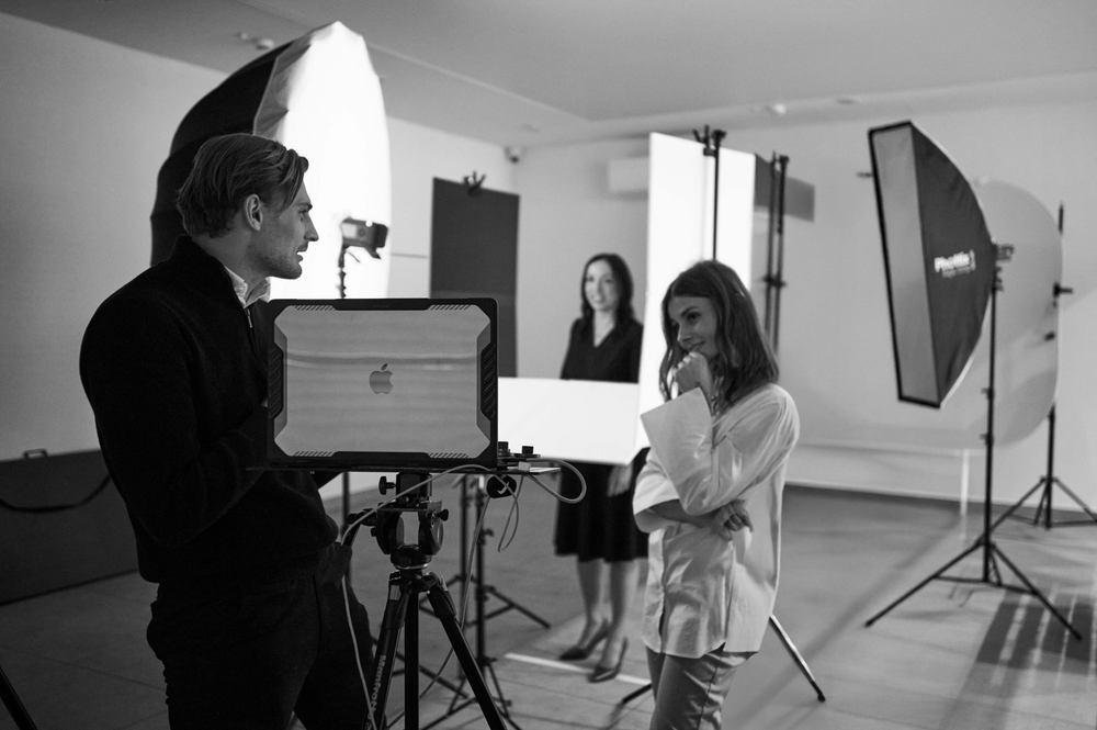 Black and white photo of a photographer directing a model during a professional studio photo shoot with lighting equipment.