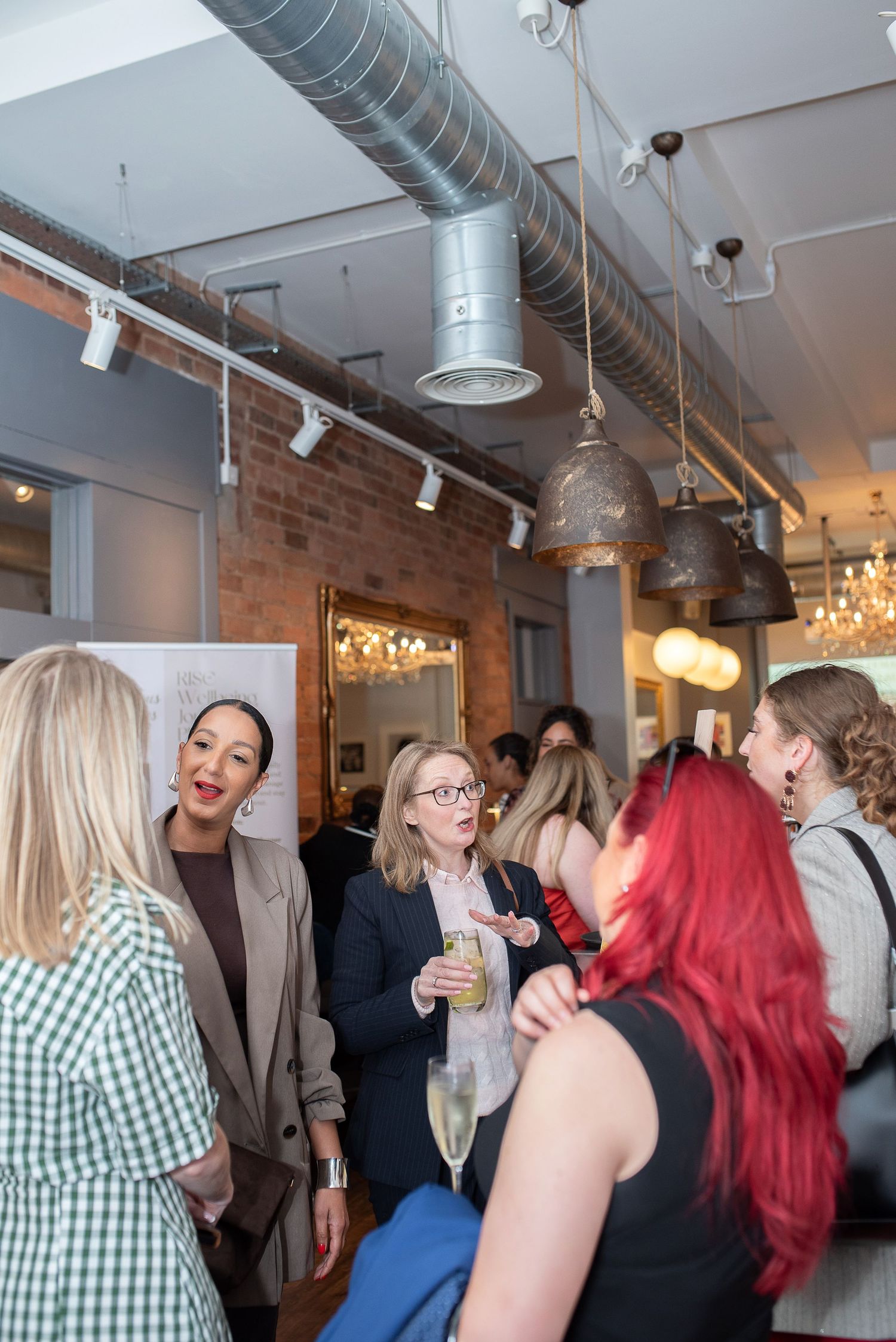 Women networking at an indoor cocktail party, holding champagne glasses in a modern industrial-style venue with exposed brick.