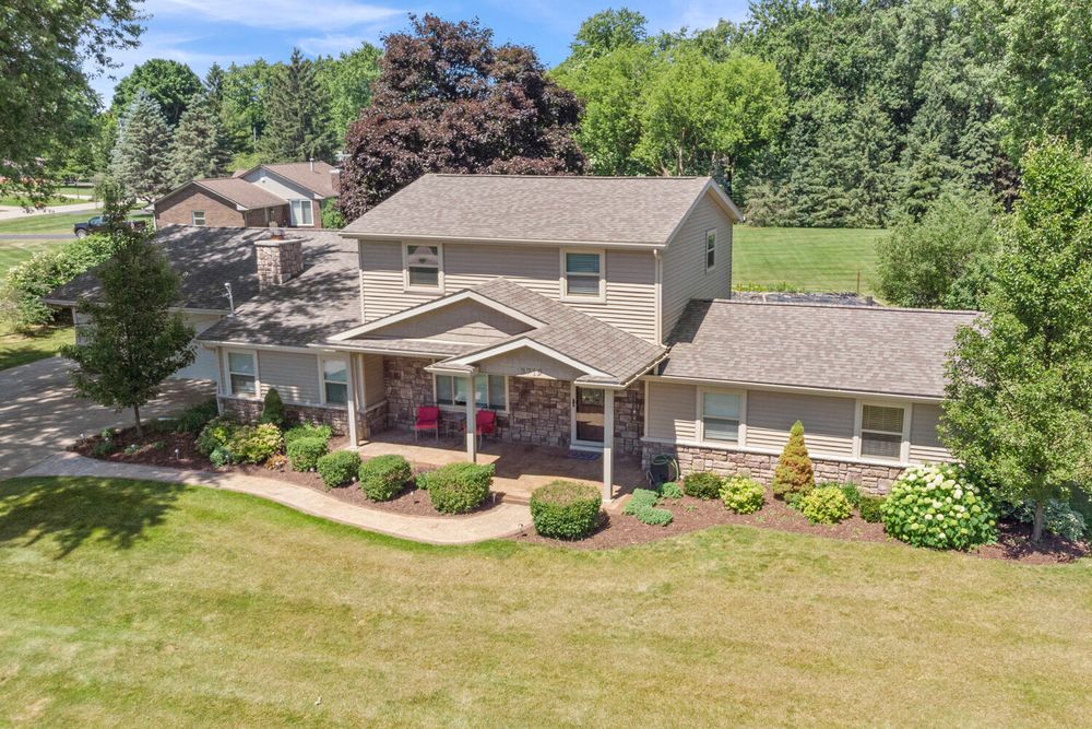 Aerial view of a two-story home with brick accents, covered porch, lush green lawn, and mature trees.