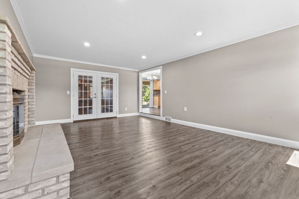 Open living room with dark hardwood floors, brick fireplace, French doors, and abundant natural light.