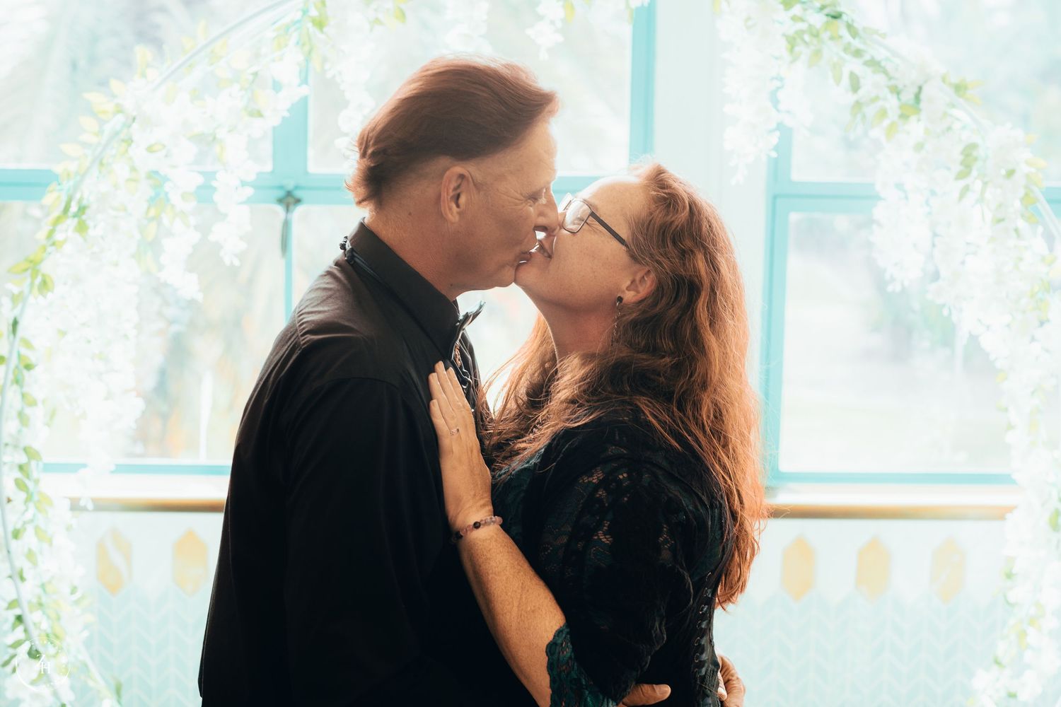 Romantic couple sharing a tender kiss under white floral decorations in a softly lit venue.