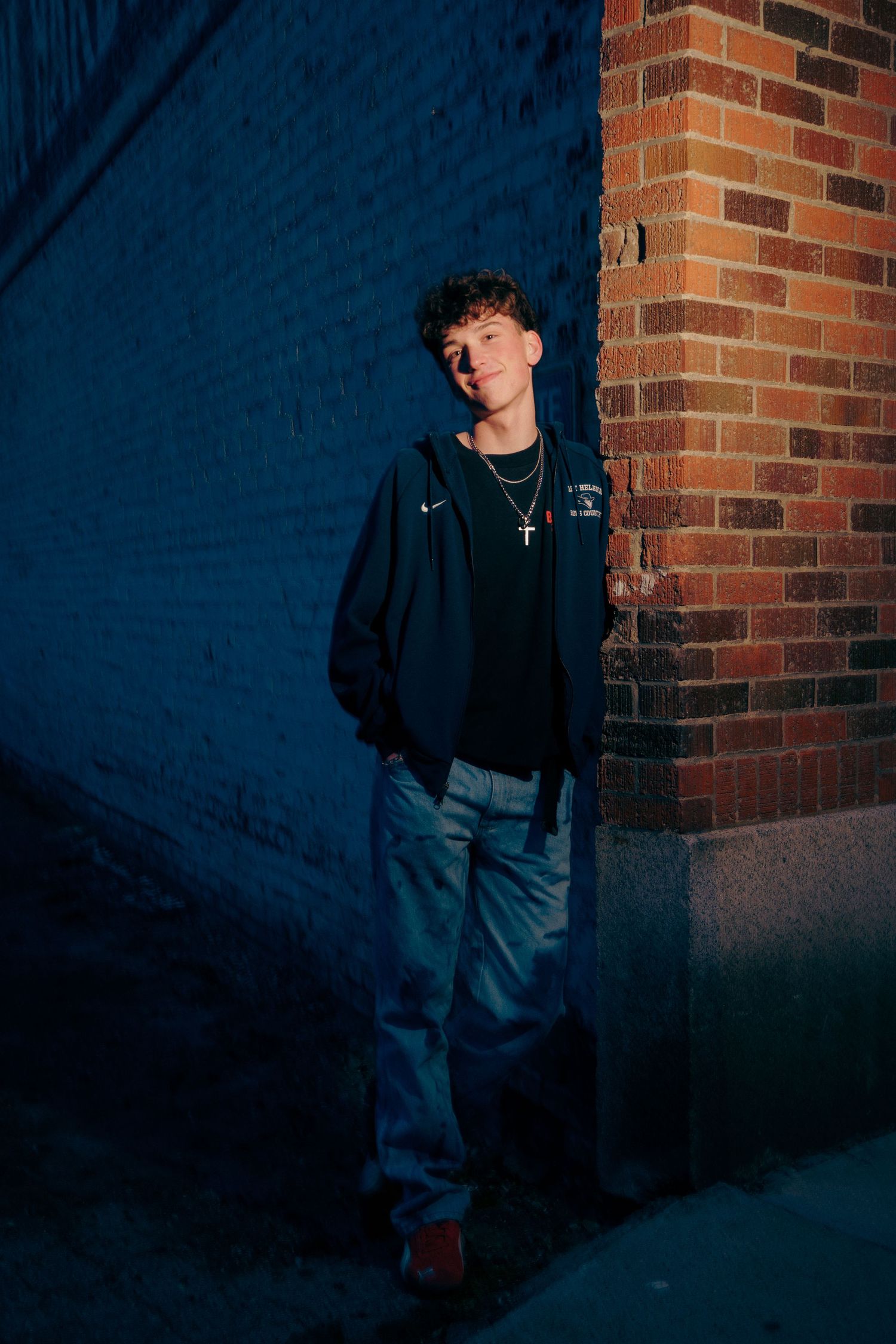 Young man in navy hoodie and jeans leaning against brick wall in dramatic blue shadow lighting.