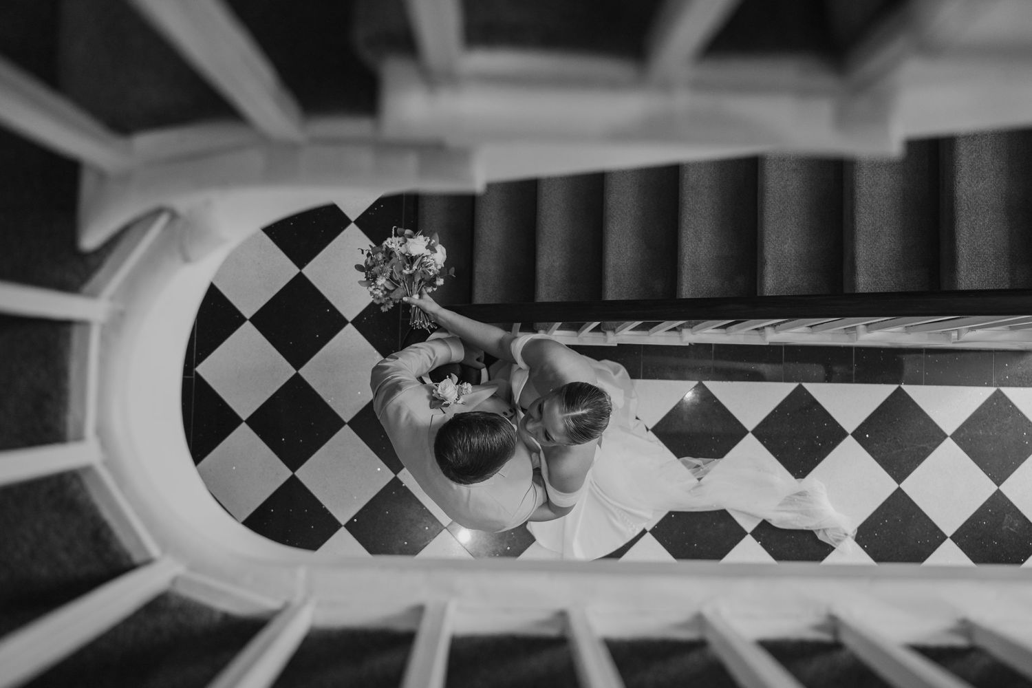Black and white aerial view of bride and groom kissing on checkered floor beneath spiral staircase.