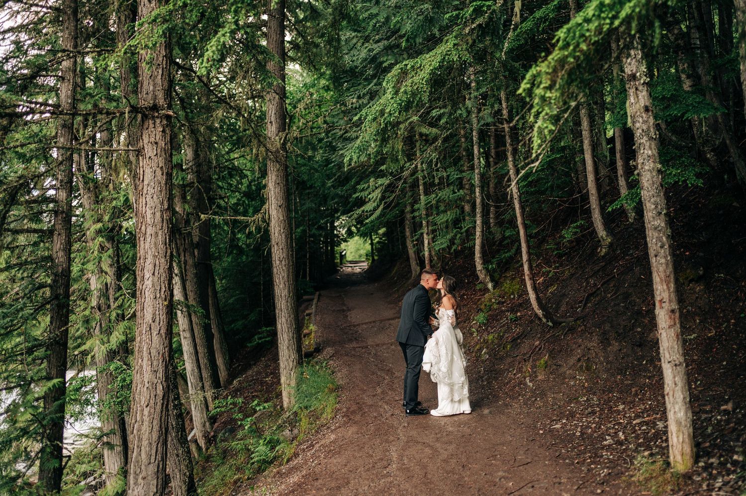 Bride and groom sharing a romantic kiss on a forest trail surrounded by tall evergreen trees.